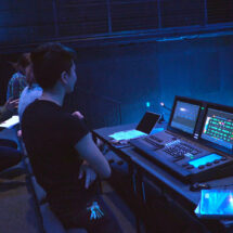 While sitting or standing near theater seats, students observe a digital lightboard and control lights on stage in a dark theater.