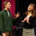 Two actors stand on a stage one with expressive face and gestures.