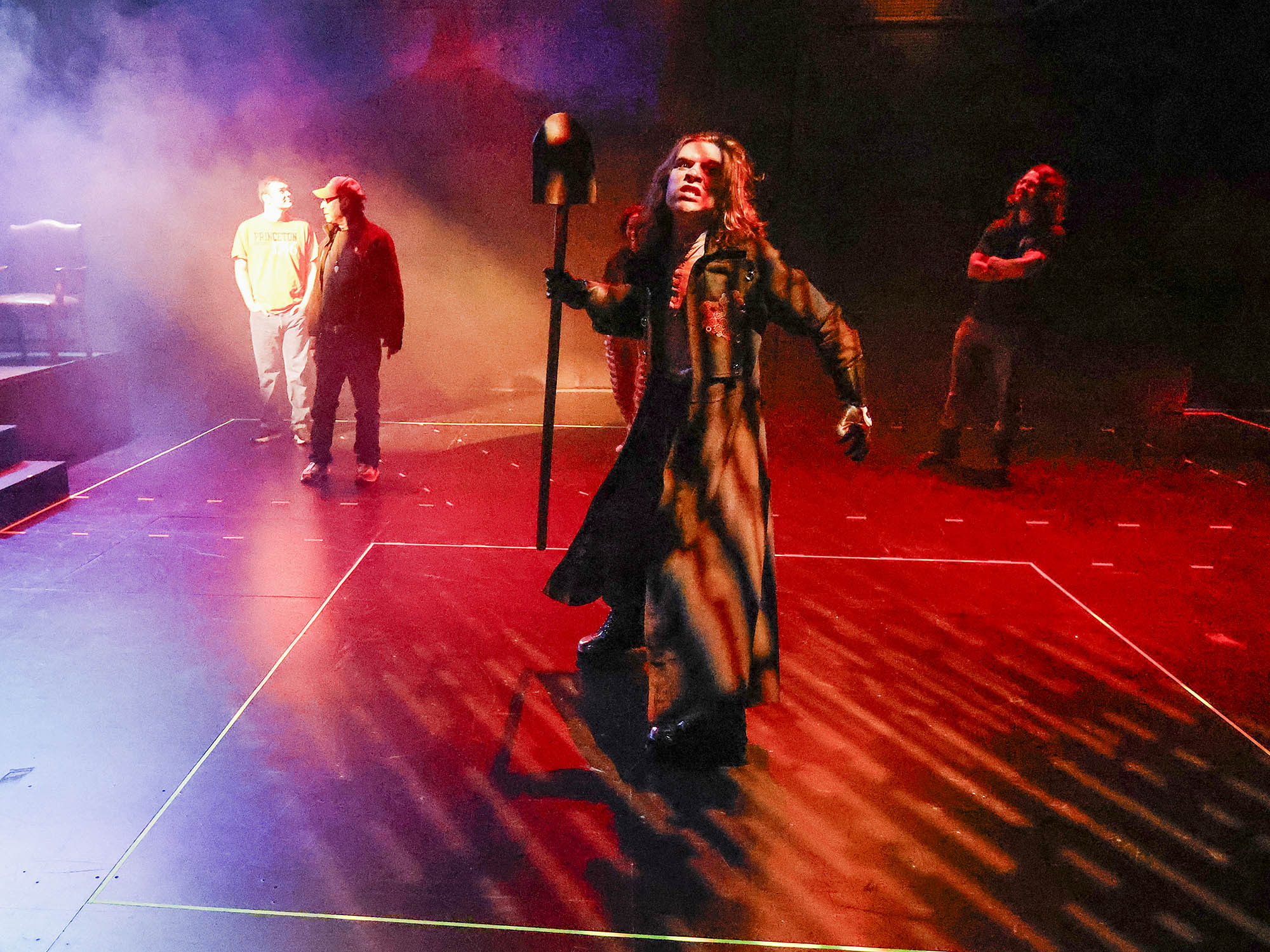 Red light and haze come from upstage as an actor in a long coat holds a shovel in a threatening manner.