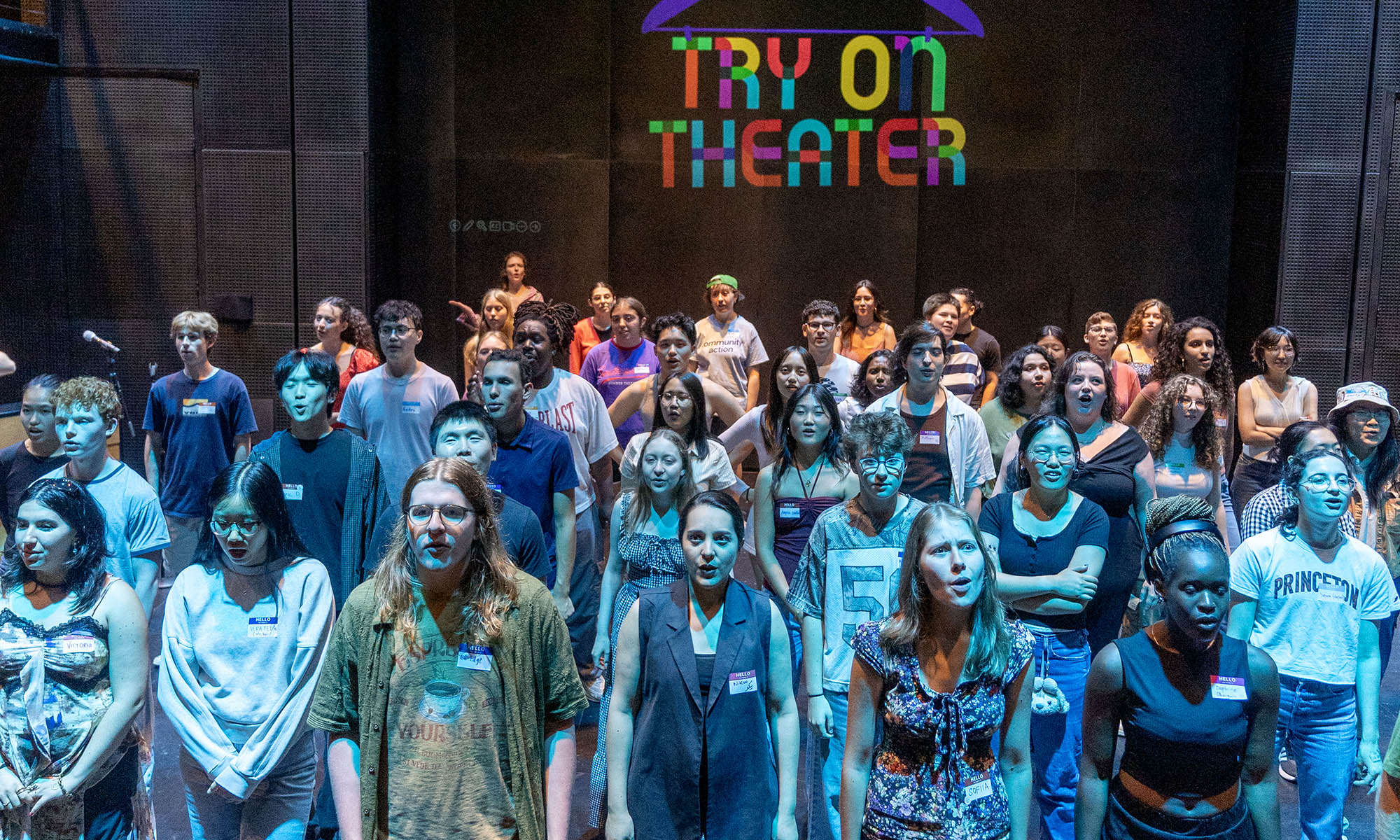 A large group of people sing on a stage with the words Try on Theater projected on the wall behind them.