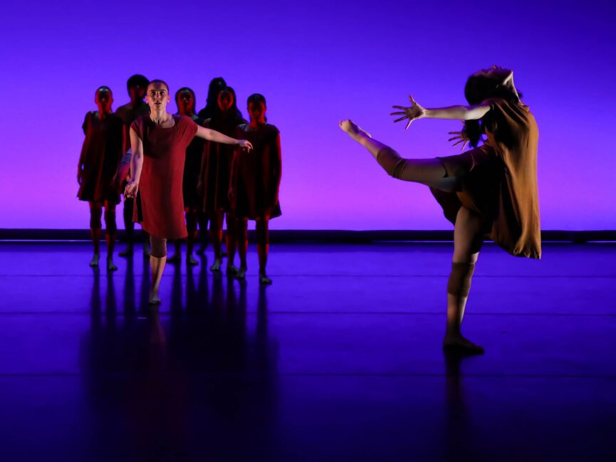 A group of dancers cluster on the left of the stage and another dancer performs solo on the right.