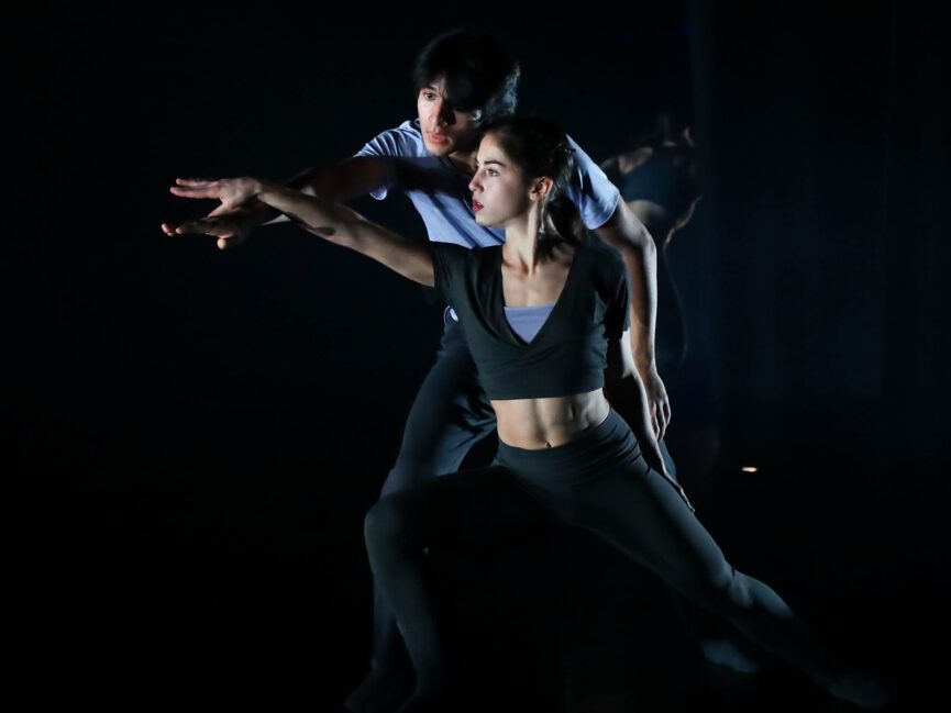 Two dancers perform on a dark stage.