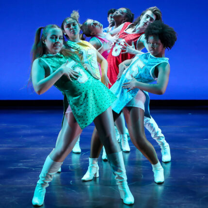 A dynamic group of dancers performing on stage, dressed in colorful, retro-inspired costumes.