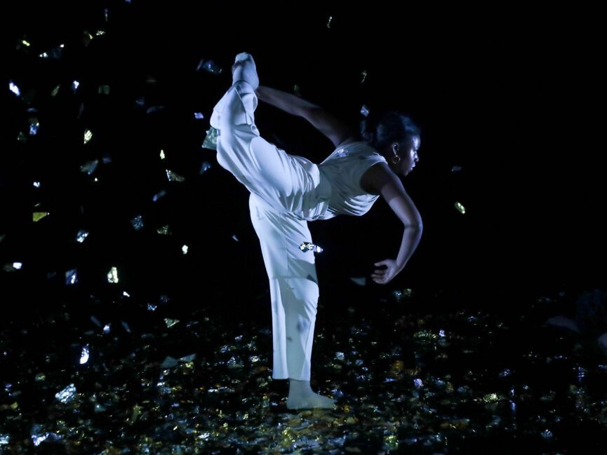 A dancer wearing a white outfit performs a high kick on a dimly lit stage scattered with reflective confetti.