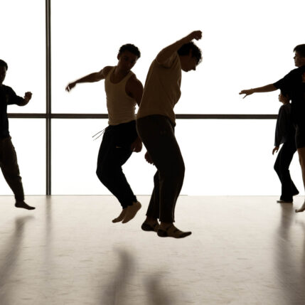A group of four dancers in various positions in front of a large window.