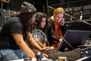 Three people work on a lighting board in a theater auditorium.