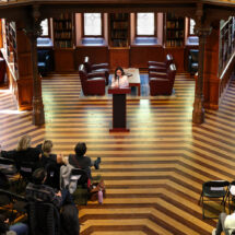 A person stands in an ornate library at a podium and reads to a crowd.