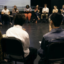 A large group of people sit in a circle in a theatrical studio setting.