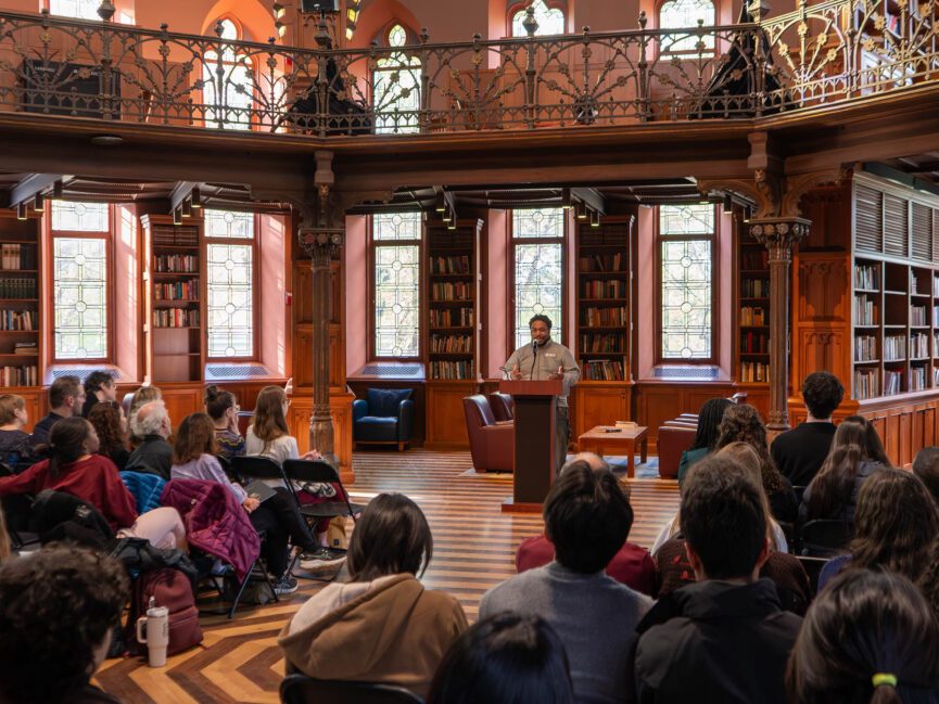 A person stands at a podium in an ornate room and reads into a microphone.