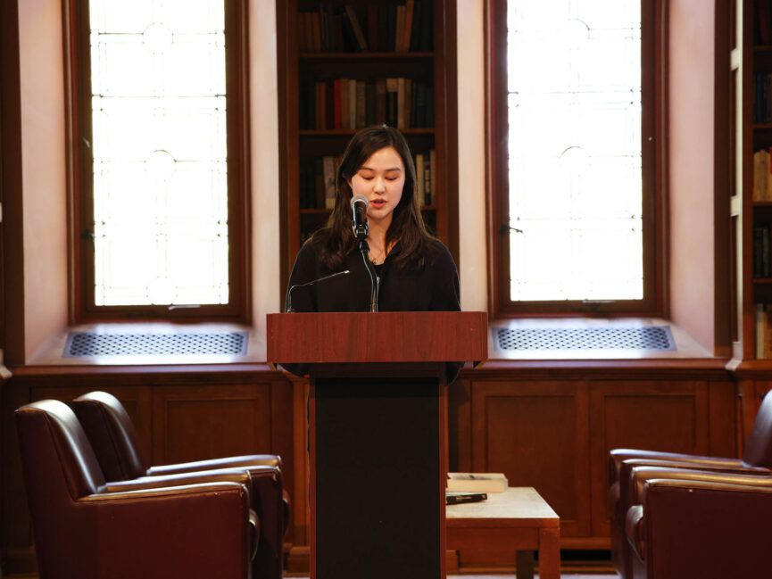 A person reads into a microphone at a podium in front of a shelf of books and two windows.