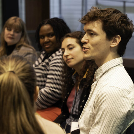 A person speaks in the foreground as others look on in the background.