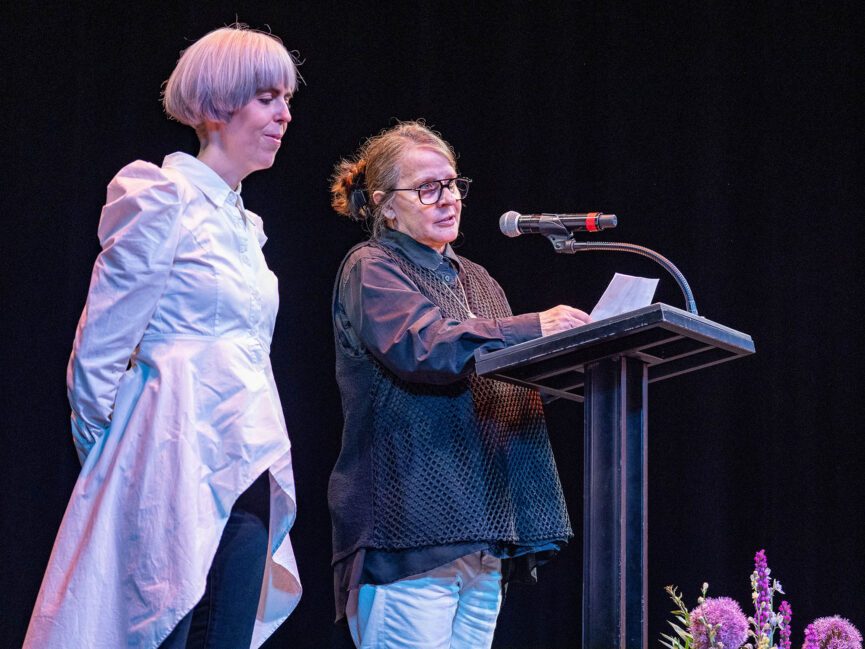 A person holds papers at a podium and speaks into a microphone as someone else stands beside them.